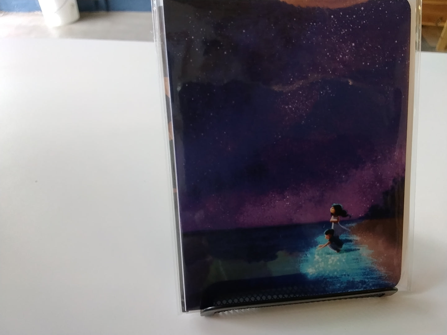 A clear plastic sleeve protects a square art print or card featuring a night sky scene with stars, purple and blue tones, and a small figure near glowing water at the bottom, photographed on a white tabletop.